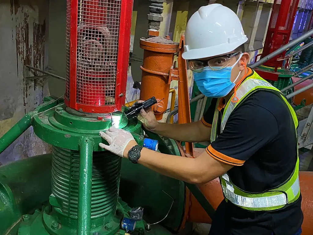 Technician wearing PPE using a flashlight to inspect a green industrial pump assembly in a mechanical plant room.