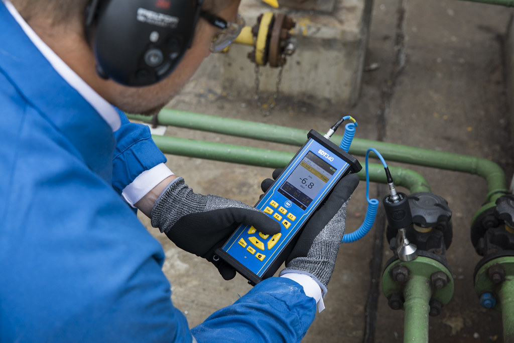 A technician wearing headphones uses an SDT340 ultrasound data collector and contact sensor to inspect industrial pipework.