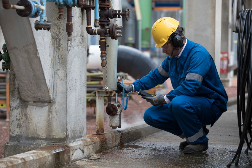 Industrial technician using a handheld ultrasound detector to inspect a valve on process piping